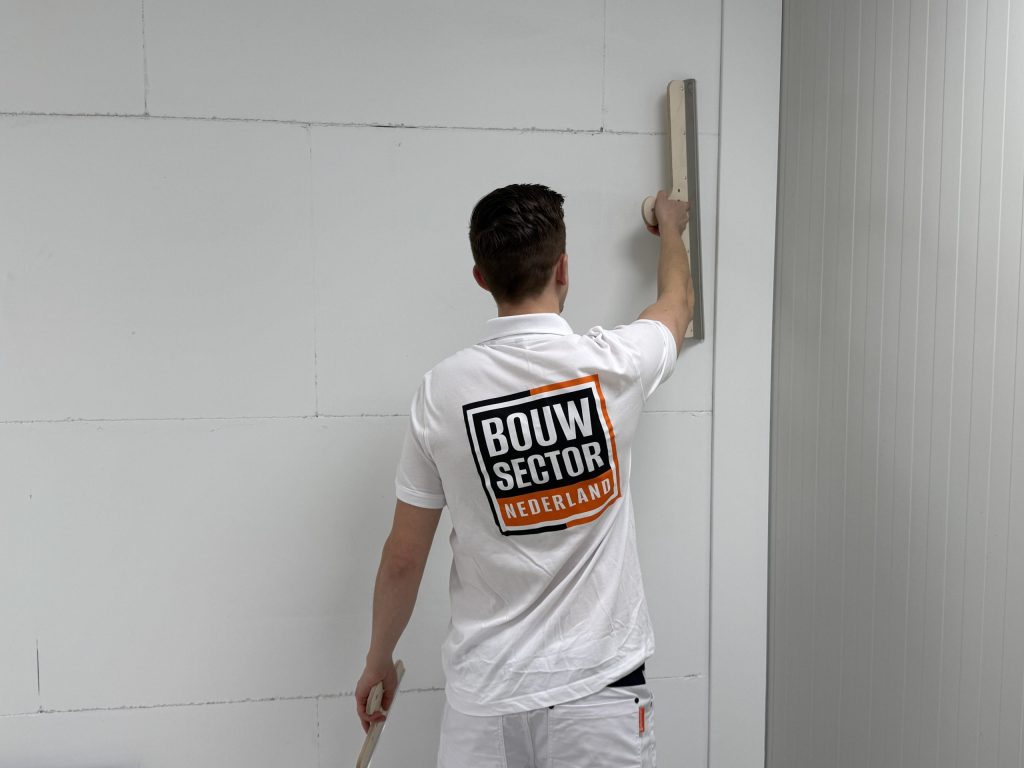 Stukadoor houdt een lange spaan hoog tegen een witte wand vlak bij de hoek van de kamer