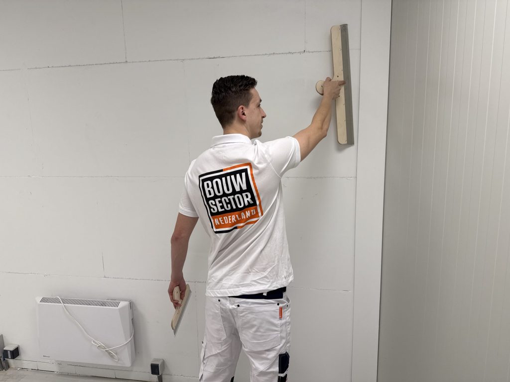 Stukadoor trekt met een lange spaan het bovenste deel van een wand vlak, naast een elektrische radiator