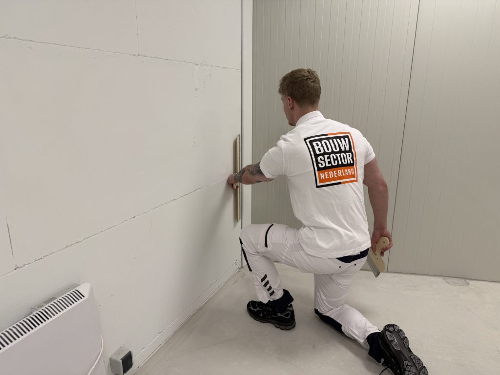 Stukadoor zit op één knie en trekt met een spaan de onderkant van een wand strak