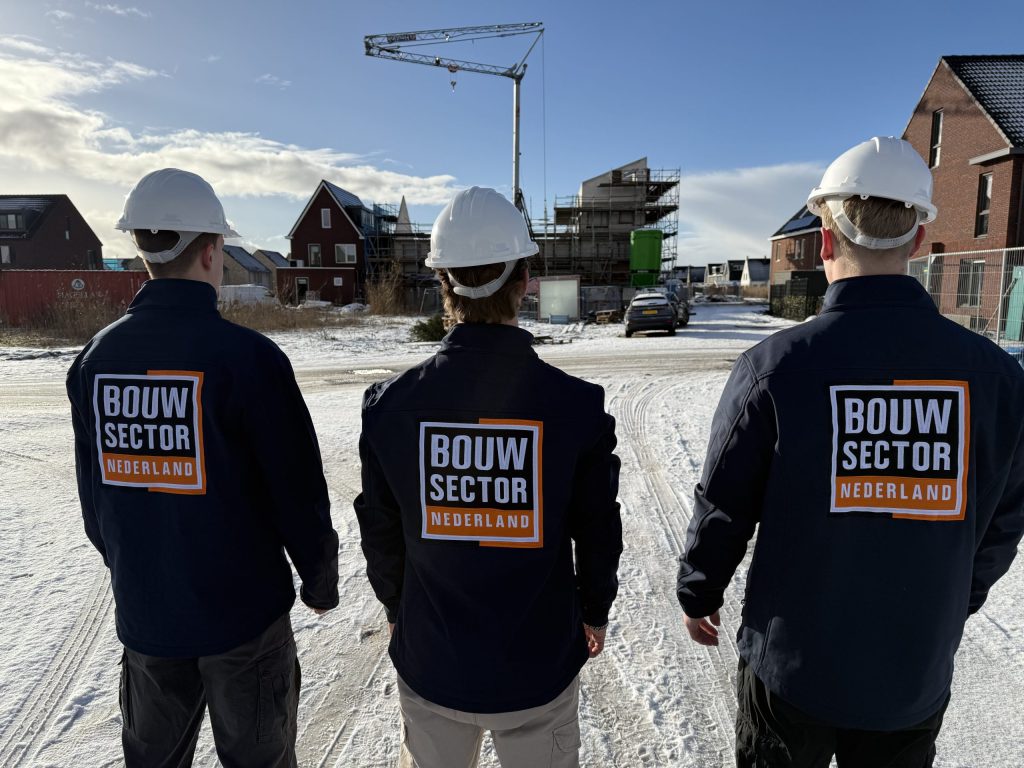 Drie vakmensen in werkjassen met logo van Bouwsector Nederland kijken naar woningen in aanbouw op een besneeuwde bouwplaats