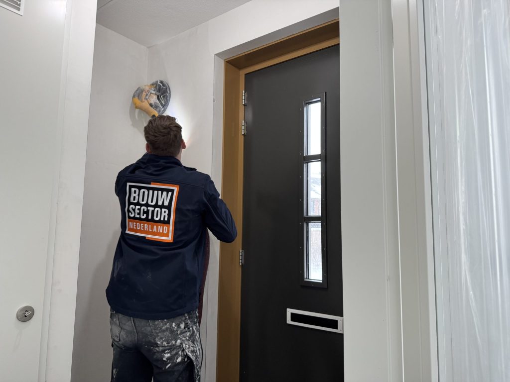 Vakman van Bouwsector Nederland in werkjas met een schuurmachine aan de bovenkant van een wand naast de voordeur